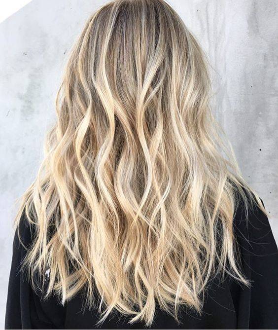Cheveux longs blonds wavy avec balayage. Coiffure blonde tendance avec reflets dorés, portés avec un pull noir. Lumière naturelle douce.
