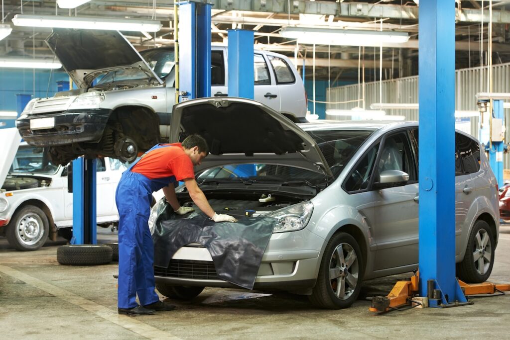 Mécanicien en bleu répare une voiture argentée dans un garage. Voitures sur élévateurs, atelier de réparation automobile.